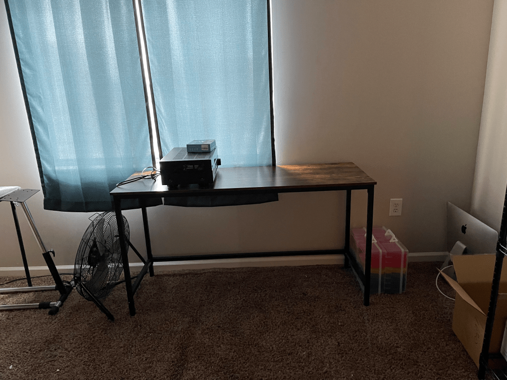 A desk in front of window with electronics on top.